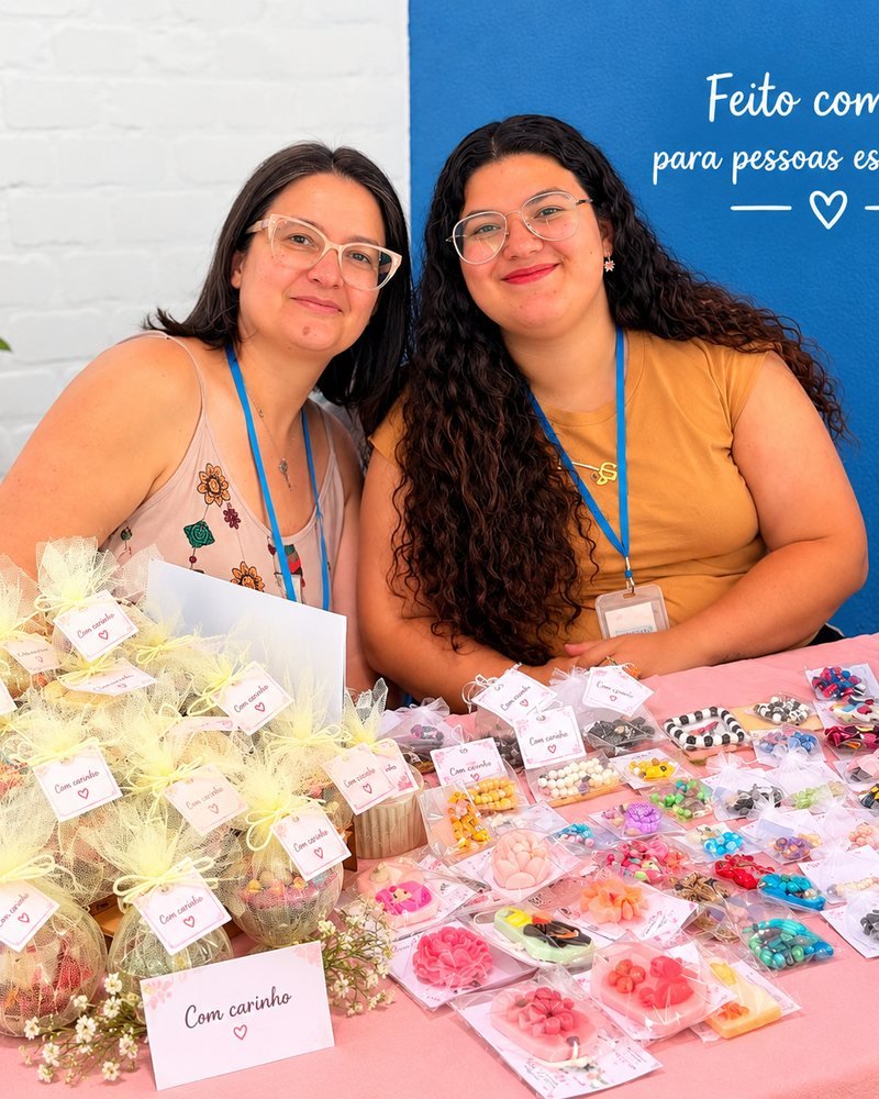 Mãe e filha, criadoras da Aura Central, sorrindo atrás da mesa com as peças artesanais