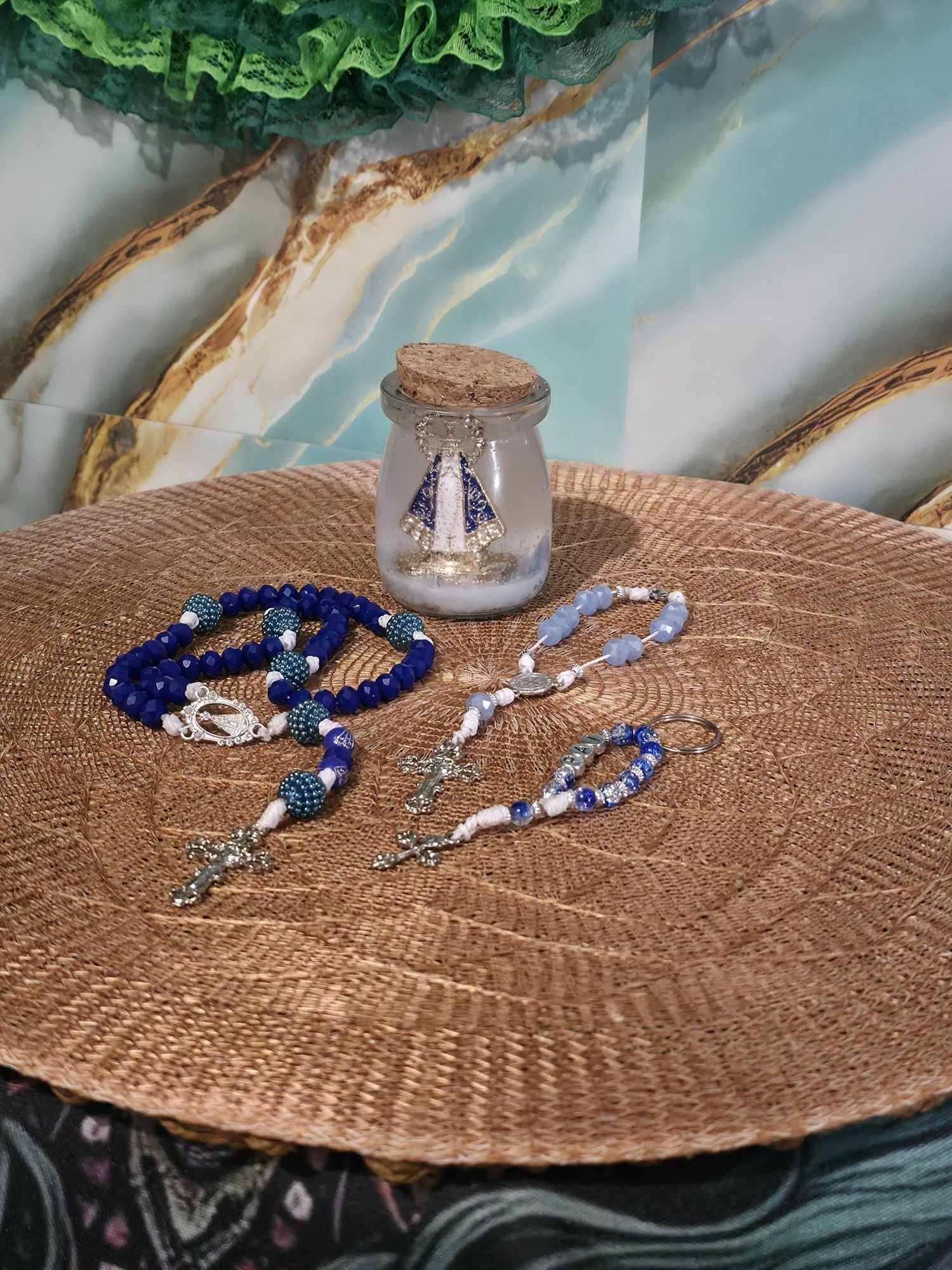 Vela aromática em pote de vidro com Nossa Senhora Aparecida e terços artesanais azuis sobre toalha de palha